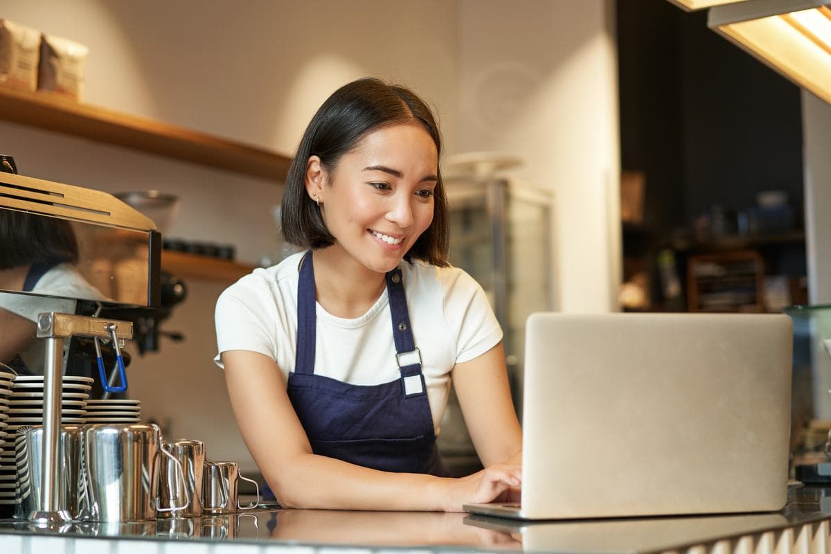 A café owner in an apron typing on a laptop behind the counter