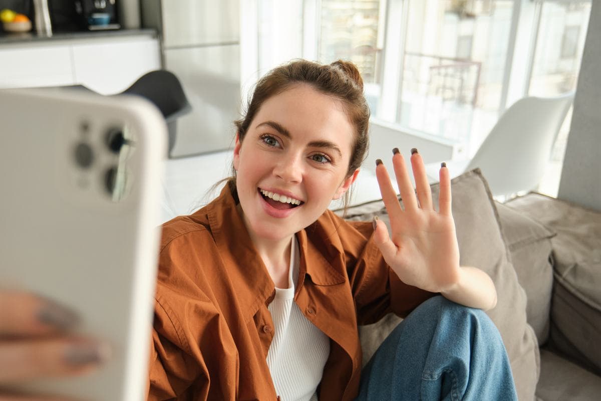 A smiling woman on a sofa waving at the phone she is recording on