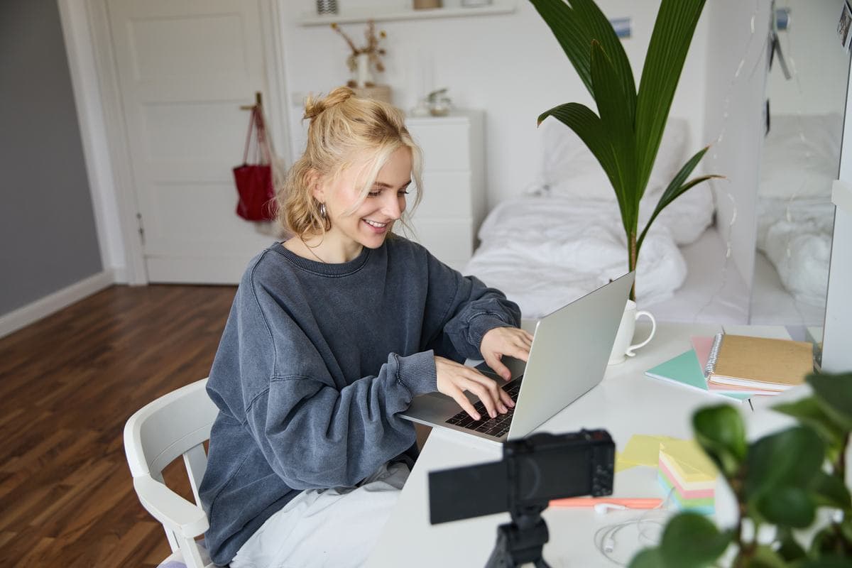 A woman at a desk recording herself with a laptop and a small camera
